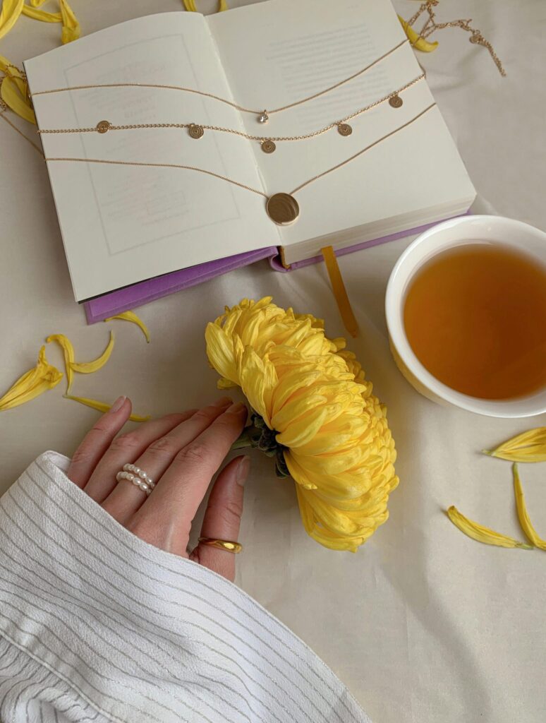 pexels photo 7664109 7664109 A serene composition featuring an open book, cup of tea, yellow flower, and hand with jewelry.